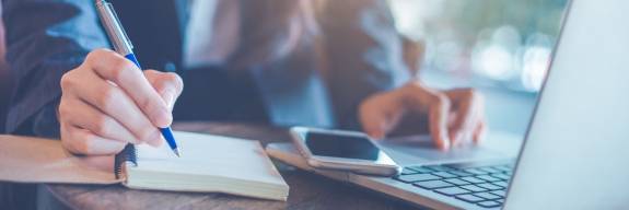 Person schreibt auf einem Notizblock mit Laptop und Smartsphone daneben